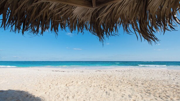 Ein Strand mit einer strohgedeckten Struktur, feinem Sand und klarem, blauem Meer.