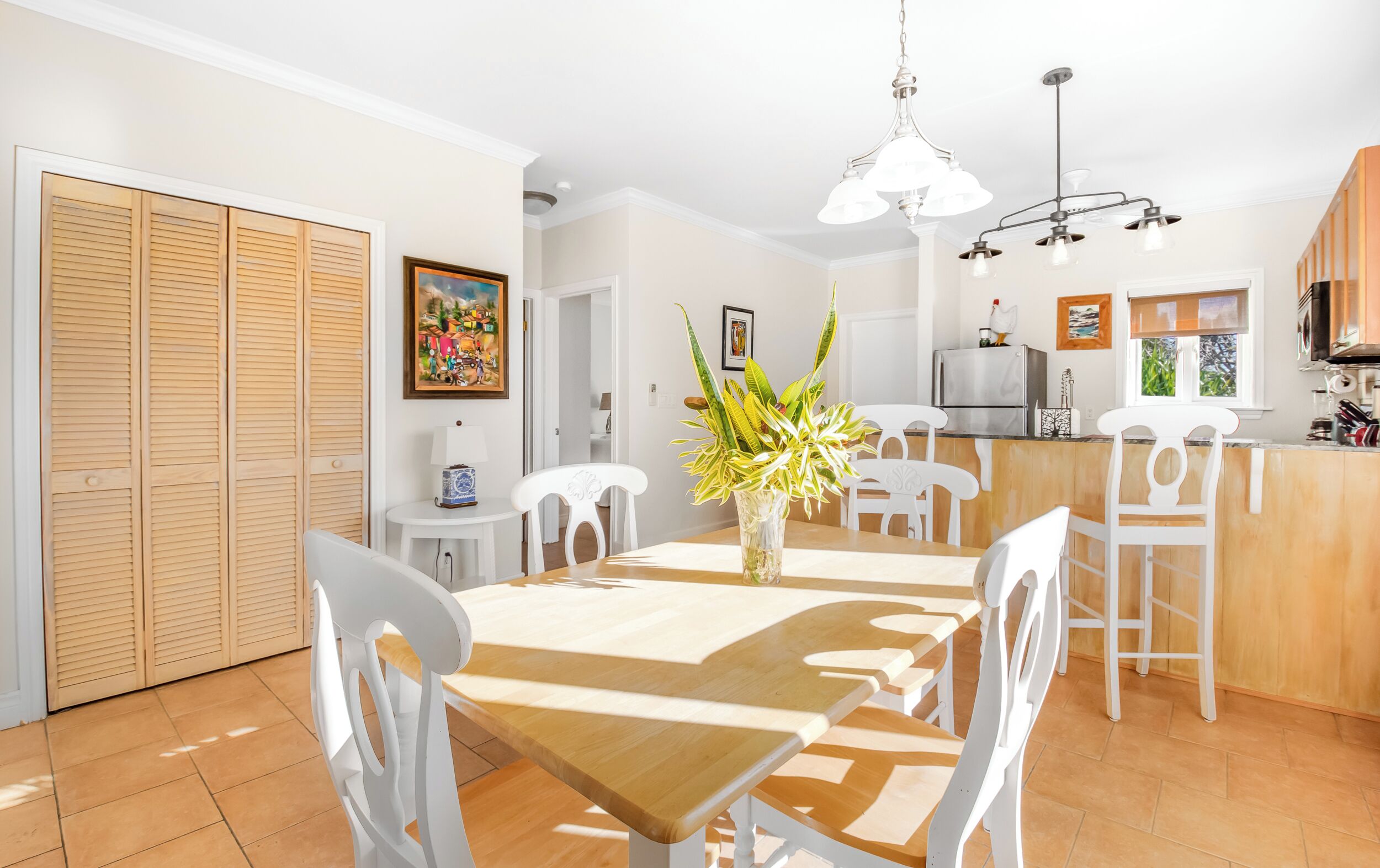 Un comedor con una mesa de madera, sillas blancas, un florero con plantas, un cuadro en la pared y una cocina al fondo.
