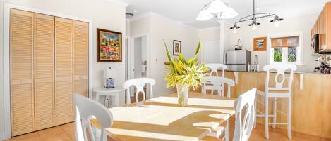 Un comedor con una mesa de madera, sillas blancas, un florero con plantas, un cuadro en la pared y una cocina al fondo.