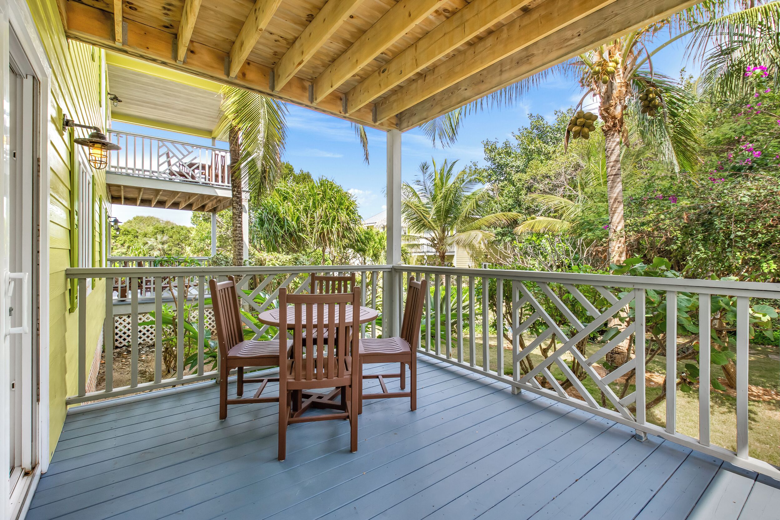 A covered outdoor area with a wooden table and chairs, surrounded by a white railing and lush greenery.