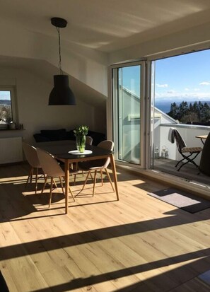 Un espace repas moderne avec une table et des chaises en bois, un luminaire suspendu et une vue sur l’extérieur grâce à de grandes portes vitrées.