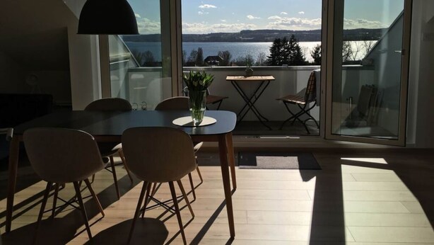 A dining area with a table and chairs, a vase with flowers, and a view of a lake through large windows. - Holiday Apartment Hammerle-seiler (Ueberlingen)