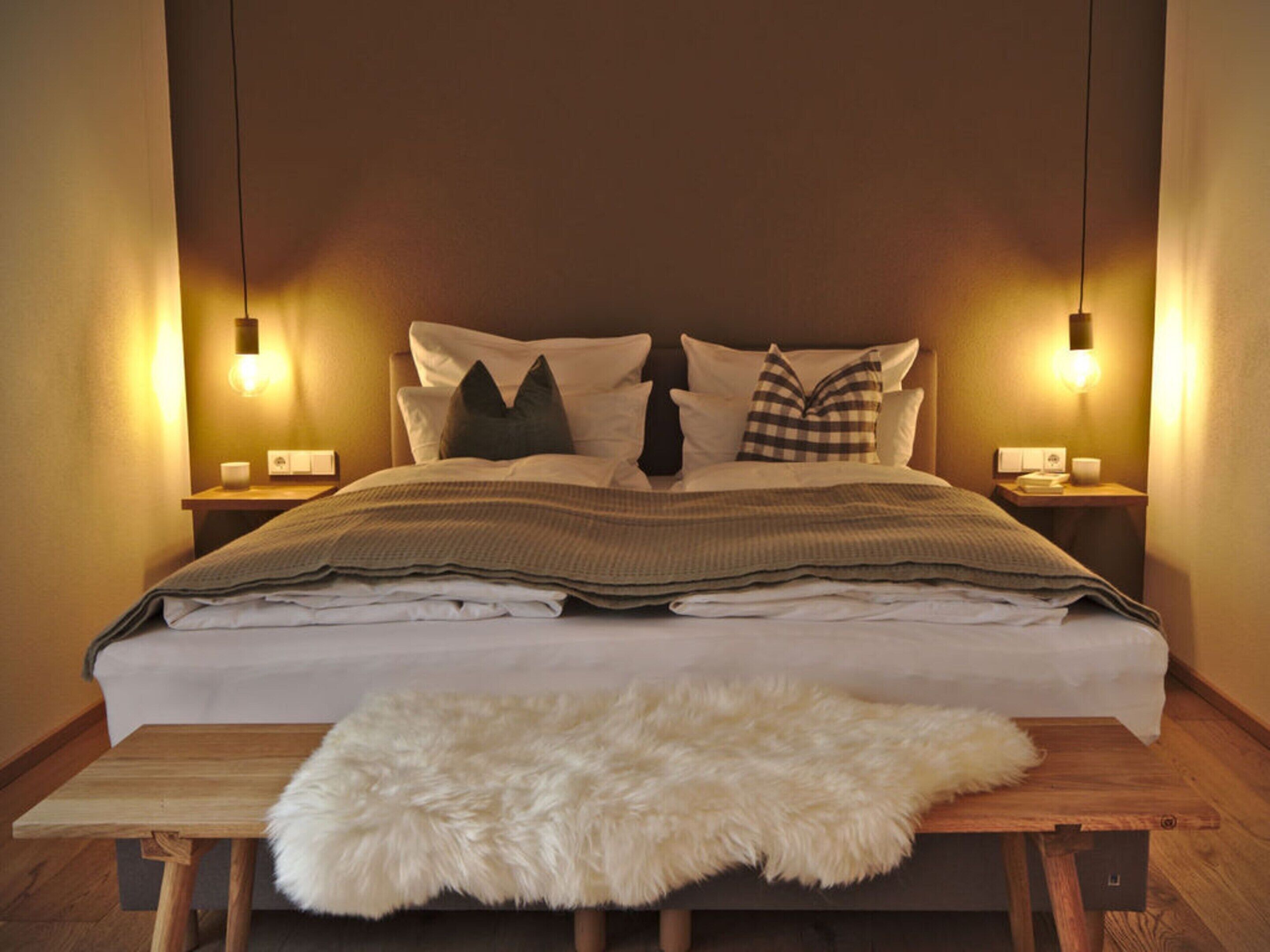 Ein ordentlich bezogenes Bett mit weißer Decke und zwei Kissen, ein flauschiger weißer Teppich auf einem Holztisch und zwei Nachttischlampen auf Nachttischen.