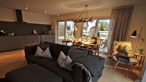 Un salon moderne avec un canapé gris foncé, un coin repas doté d’une table en bois et une vue sur l’extérieur grâce à de grandes fenêtres.