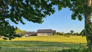 Une grande maison de plain-pied, dotée d’un toit en pente, entourée d’un champ herbeux et d’arbres.