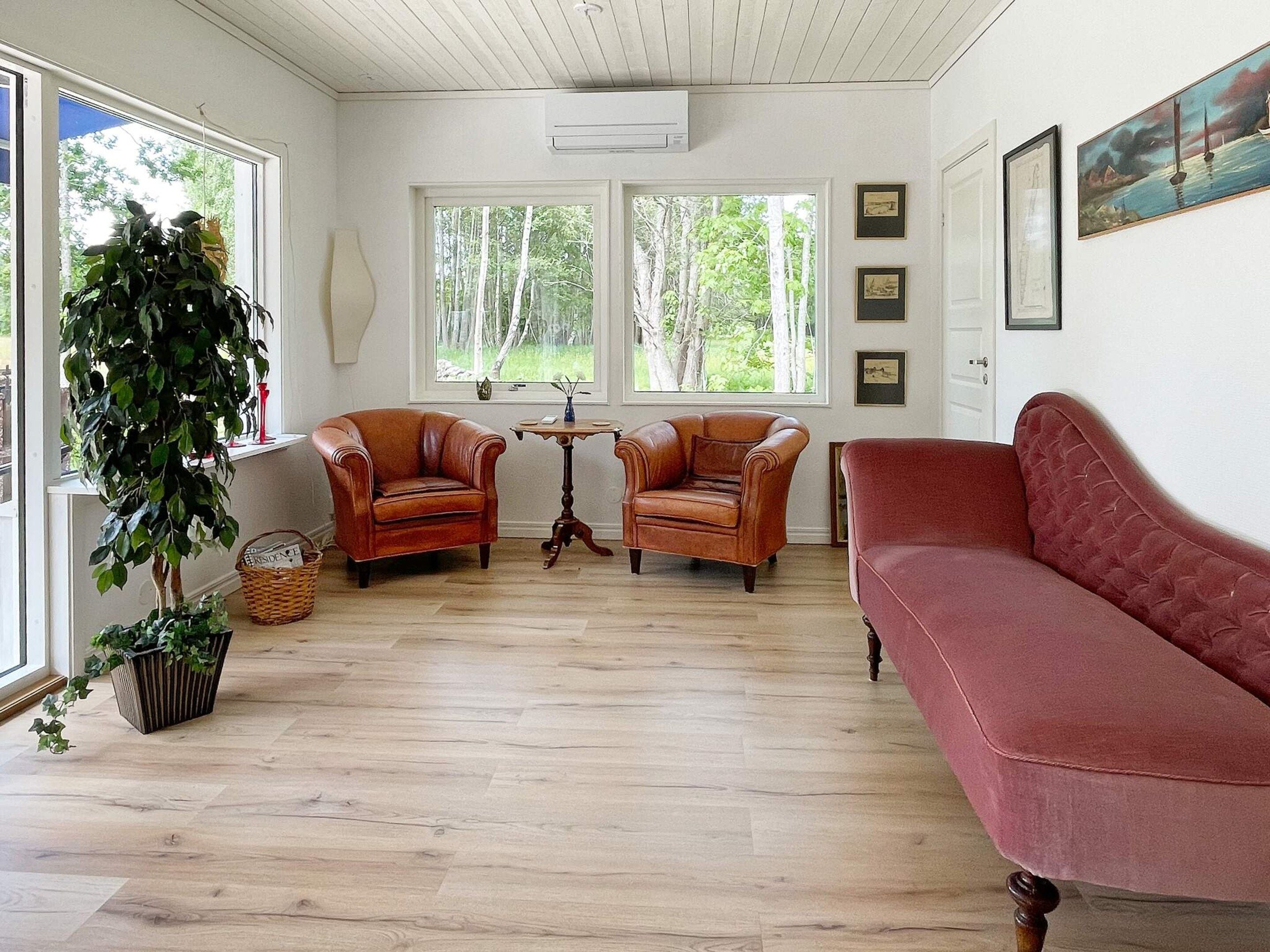 A room with two leather armchairs, a red sofa, a wooden floor, and a large window with a view of trees.
