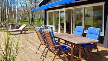 Una terraza exterior con una mesa de madera y sillas azules, una silla de lounge y un vidrio que da a un área cubierta.