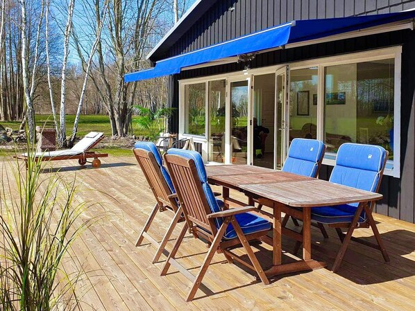 An outdoor deck with a wooden table and blue chairs, a lounge chair, and a glass door leading to a covered area. - 4 Star Holiday Home in Lottorp (Löttorp)