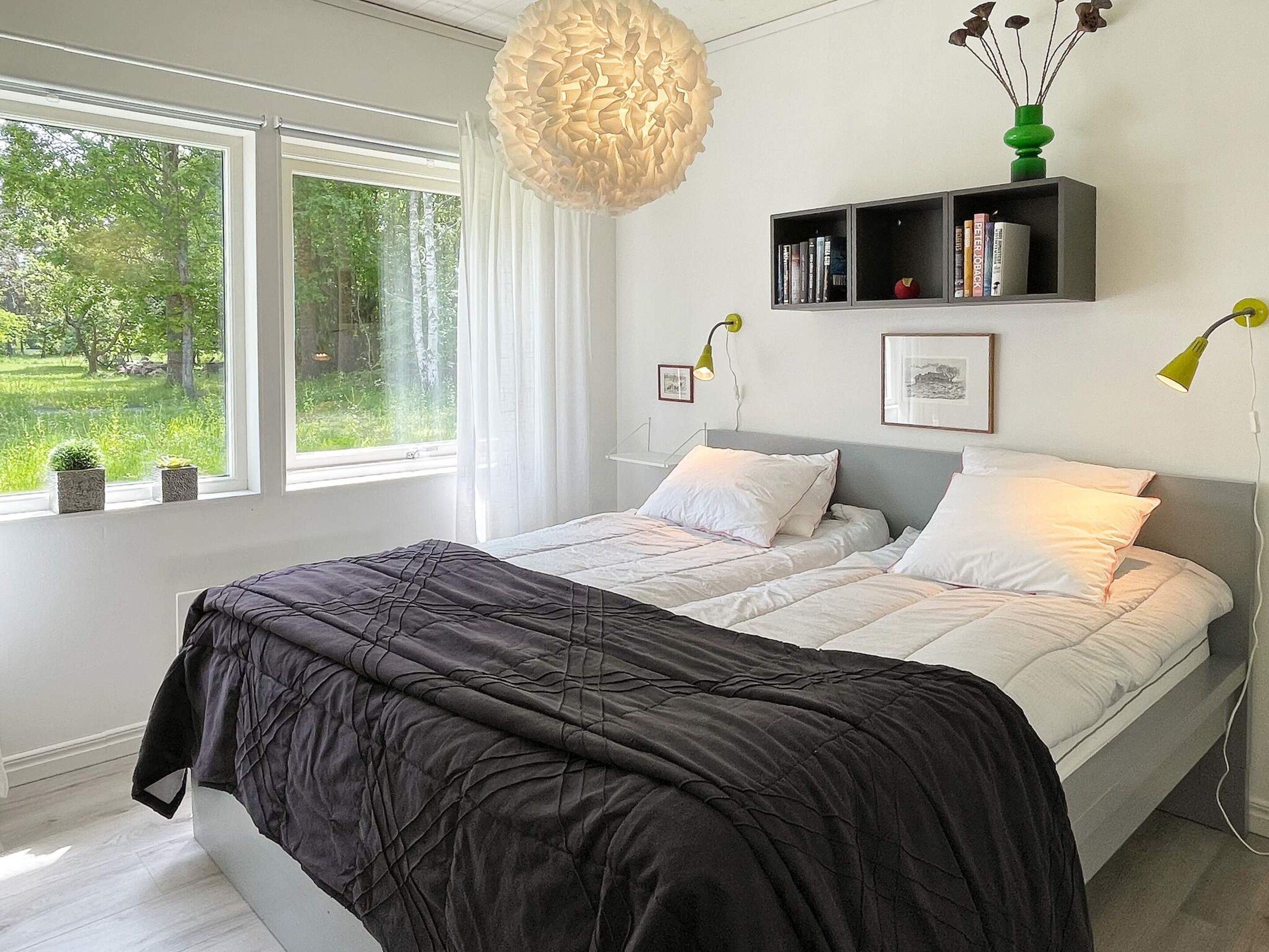 Un dormitorio con cama, una mesita de noche, una ventana con cortinas, una repisa en la pared con libros y una lámpara colgante.