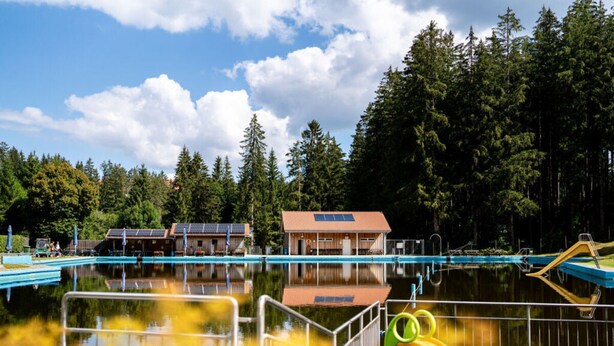 A swimming pool with a slide, surrounded by trees and a building with solar panels. - History and Luxus (Schonach im Schwarzwald)