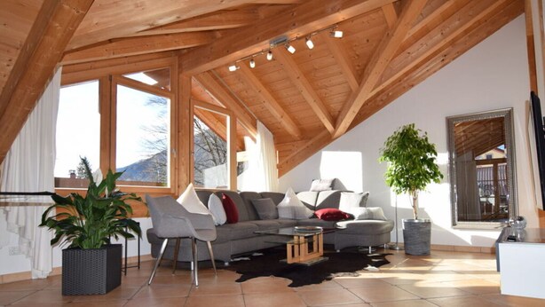 A spacious loft with a high ceiling, wooden beams, a grey sofa set, a coffee table, a potted plant, and a large mirror. - Pleasant Holiday Home With View (Garmisch-Partenkirchen)
