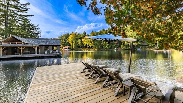 Een houten terras met lounge stoelen en een tafel, met uitzicht op een rustig meer met een blokhut en een steiger op de achtergrond.