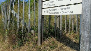 En vejskilteviser med pile, der peger mod Strand - Plage, Strand, og Bewaakt - Surveillé Bewaachtet - Bevogtet.