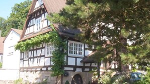 A traditional half-timbered house with a steeply pitched roof, a stone archway entrance, and a small garden area with a potted plant. - Pfaelzerleben (Oberotterbach)