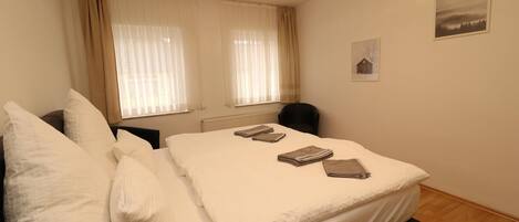 A neatly made bed with white linens and a few folded towels on top. There is a chair next to the bed and a window with sheer curtains.