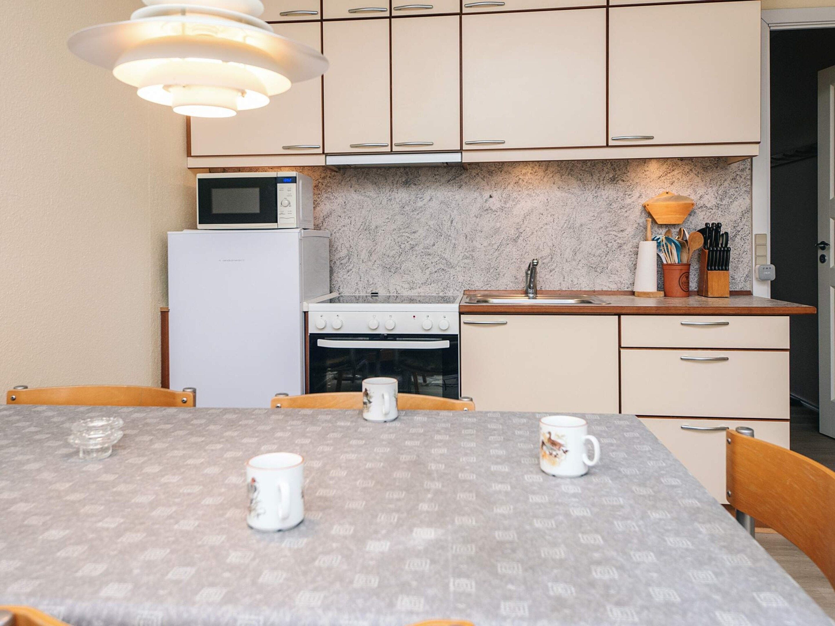 Une cuisine compacte équipée d’un four à micro-ondes, d’une cuisinière, d’un four et d’un évier. On y trouve une table avec deux tasses et une chaise.