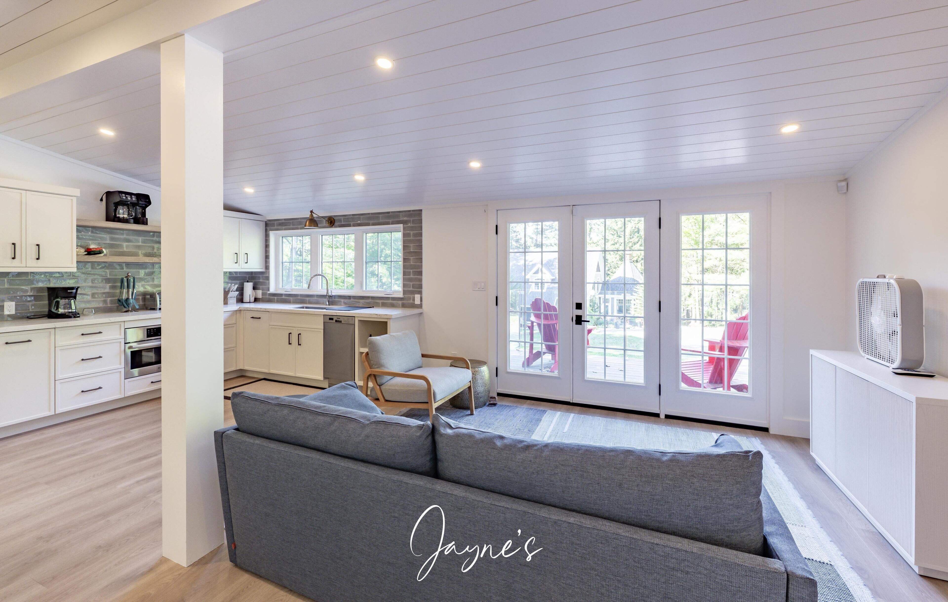 A modern kitchen with white cabinets, a built-in oven, and a coffee machine. There is a grey sofa in the living area with a view of the outside.