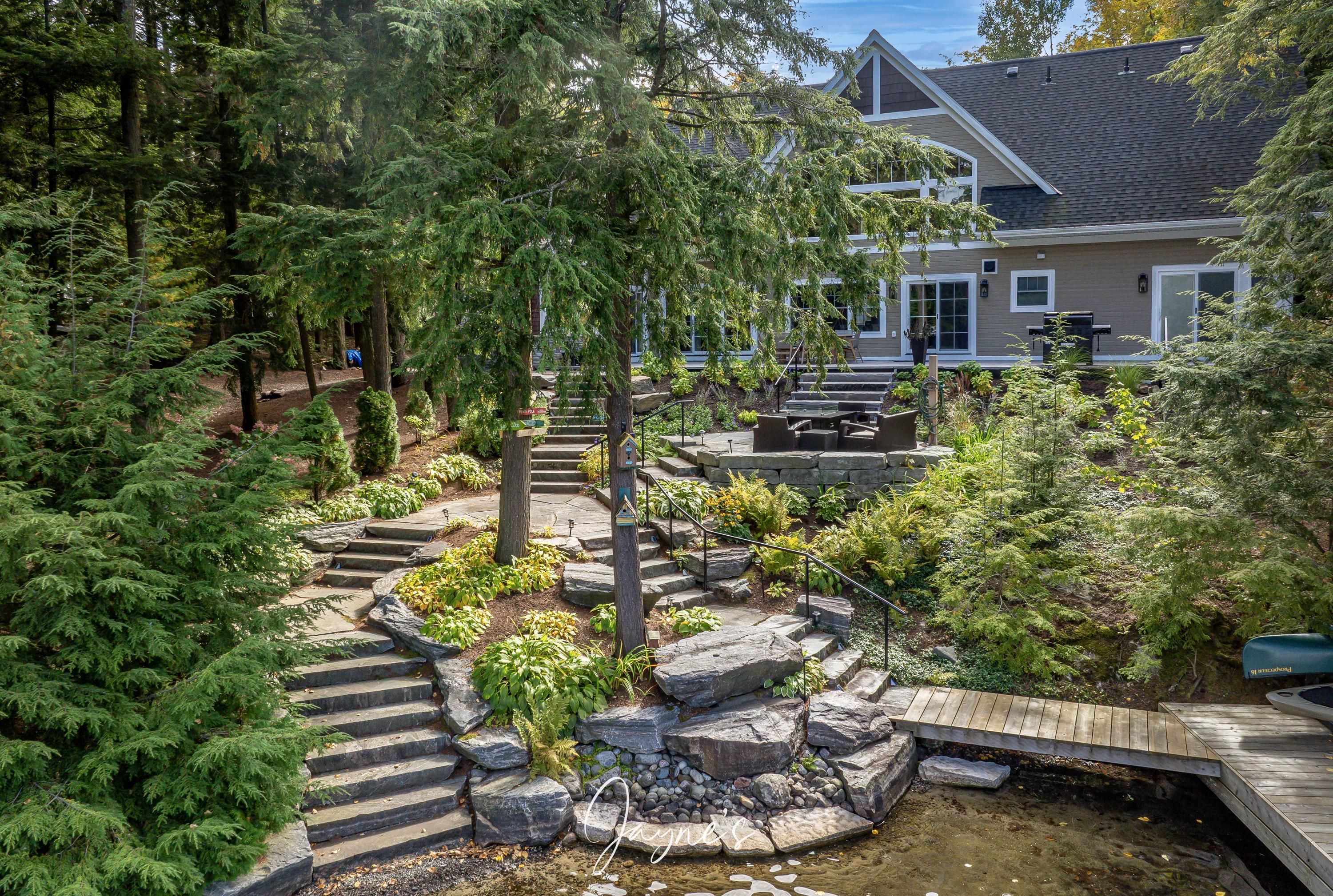 A residential property with a landscaped garden, a wooden deck, and a stone staircase leading to a house with a grey roof.