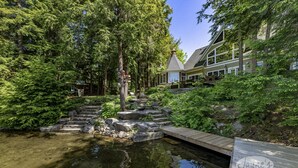 A pond with stone steps leading to a wooden dock, surrounded by dense greenery and a building with a gabled roof. - 7 bed, 5 bath spectacular cottage on Lake Joseph! (Parry Sound Mid-Shore (Bala))