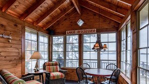 L’intérieur chaleureux d’un chalet, avec une grande fenêtre donnant sur le lac Muskoka, un fauteuil en osier, une petite table avec une lampe et un plafond en bois.