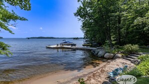 Une jetée au bord du lac, avec un bateau amarré, entourée d’arbres et bordée d’une plage de sable où sont disposés des chaises longues.