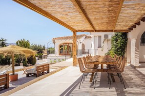 Une terrasse extérieure avec une table et des chaises en bois, une pergola et une vue sur un bâtiment blanc orné d’arcades.