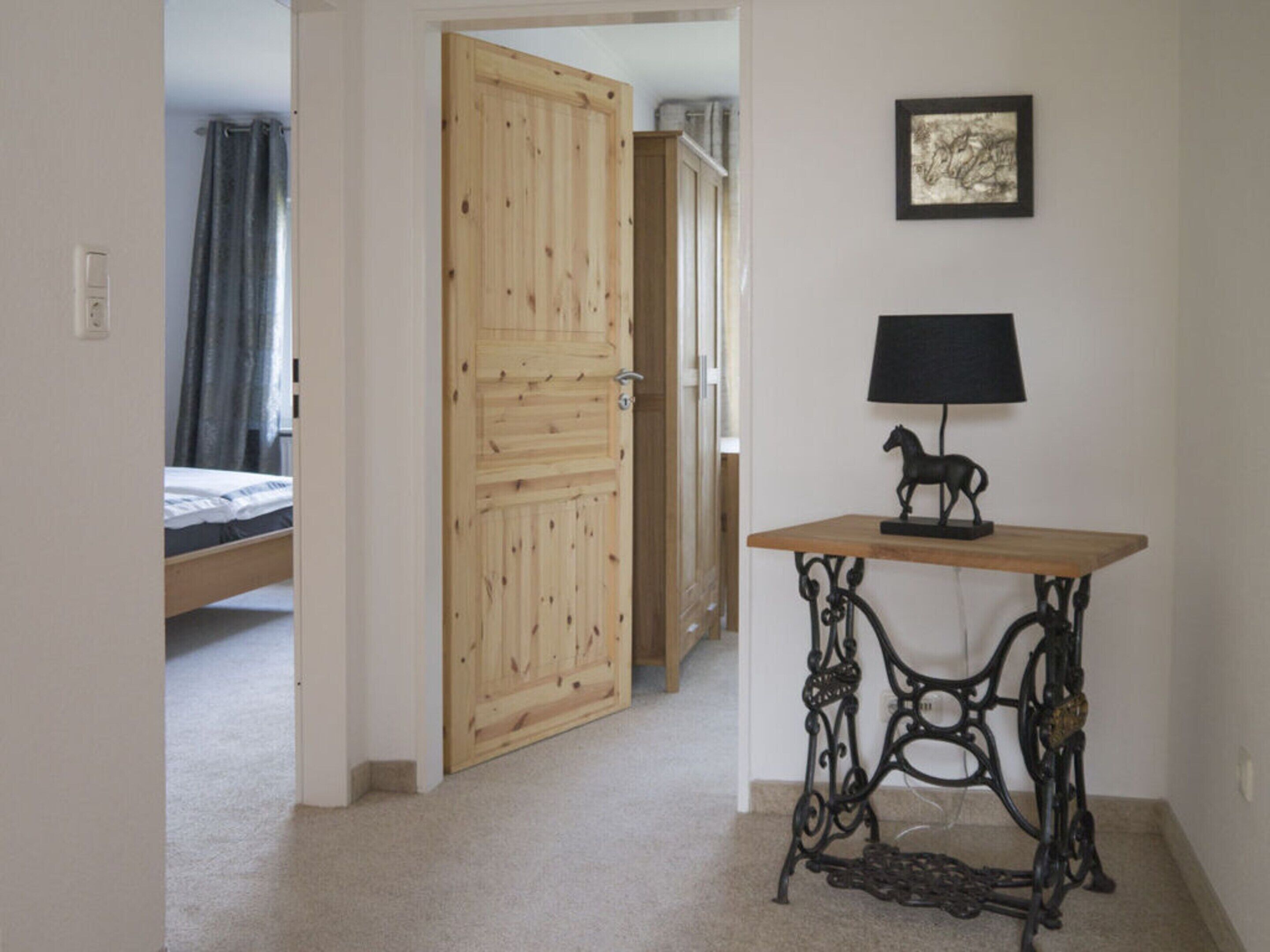 A bedroom with a wooden door, a bed, a small table with a lamp, and a framed picture on the wall.