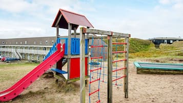 A playground with a slide and climbing structures, set against a backdrop of a building and sandy terrain.