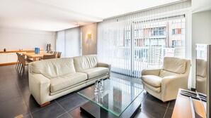 A modern living room with a sofa, armchair, and glass coffee table. There is a dining area with chairs and a table, and large windows with blinds. - Apartment in Belgium 100m From the Sea (Koksijde)