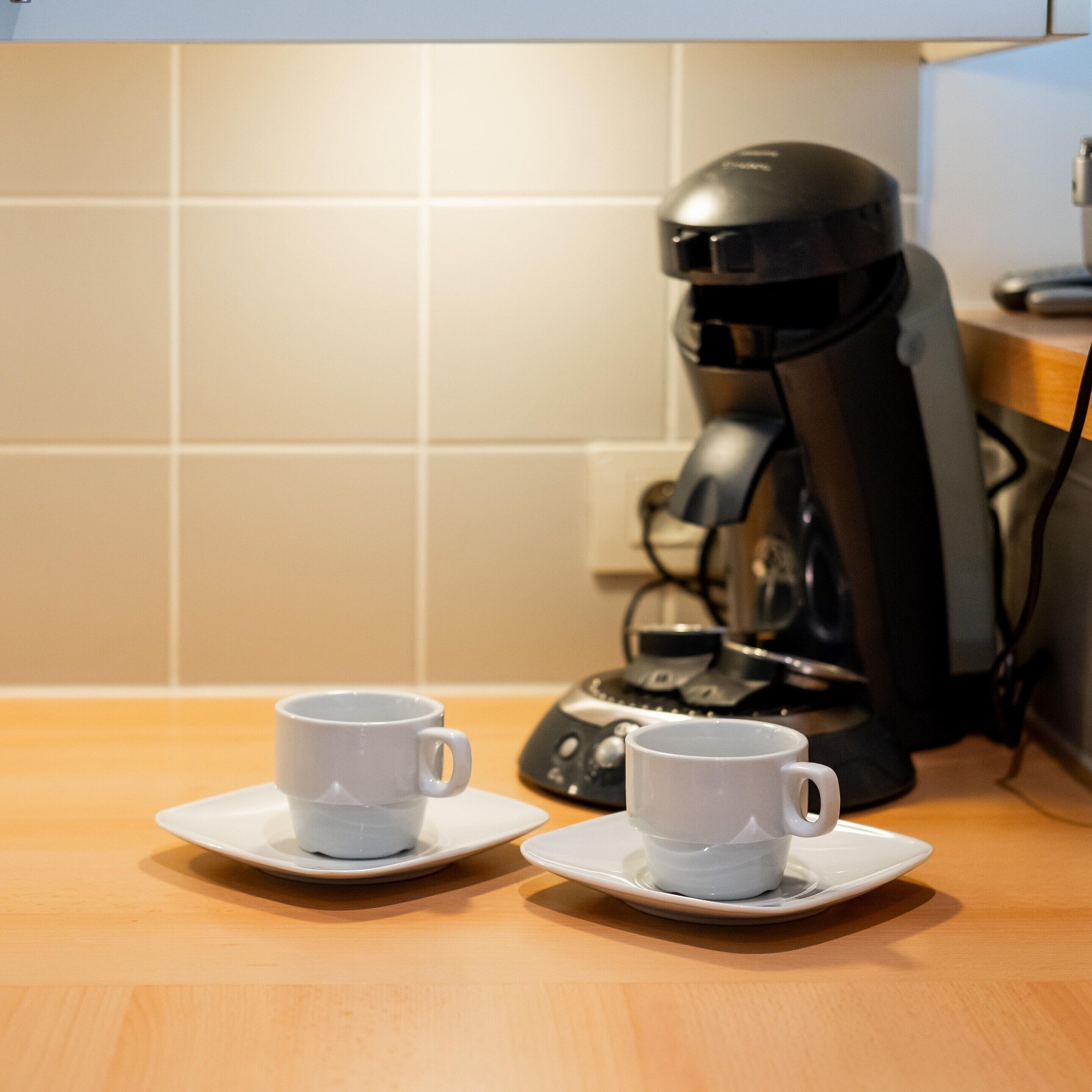 Twee witte kopjes op schoteltjes op een houten tafel, met een koffiezetapparaat op de achtergrond.