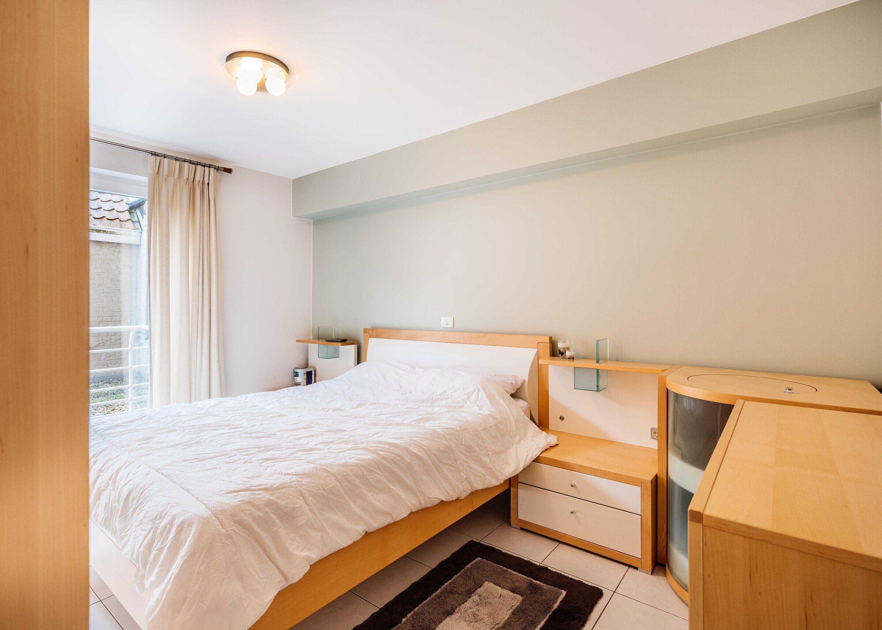 Quarto com cama, mesas de cabeceira, cômoda e janela com cortinas.