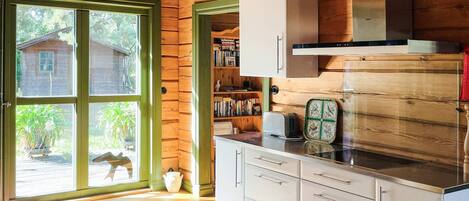 Ein Cabin-Innenraum mit Holzfassade, einer Küchenzeile mit weißen Schränken, einer eingebauten Bücherwand und einem großen Fenster mit Blick auf ein Holzhaus und viel Grün.