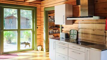 Interior de una cabaña de madera con zona de cocina, una estantería y vistas a través de un ventanal.