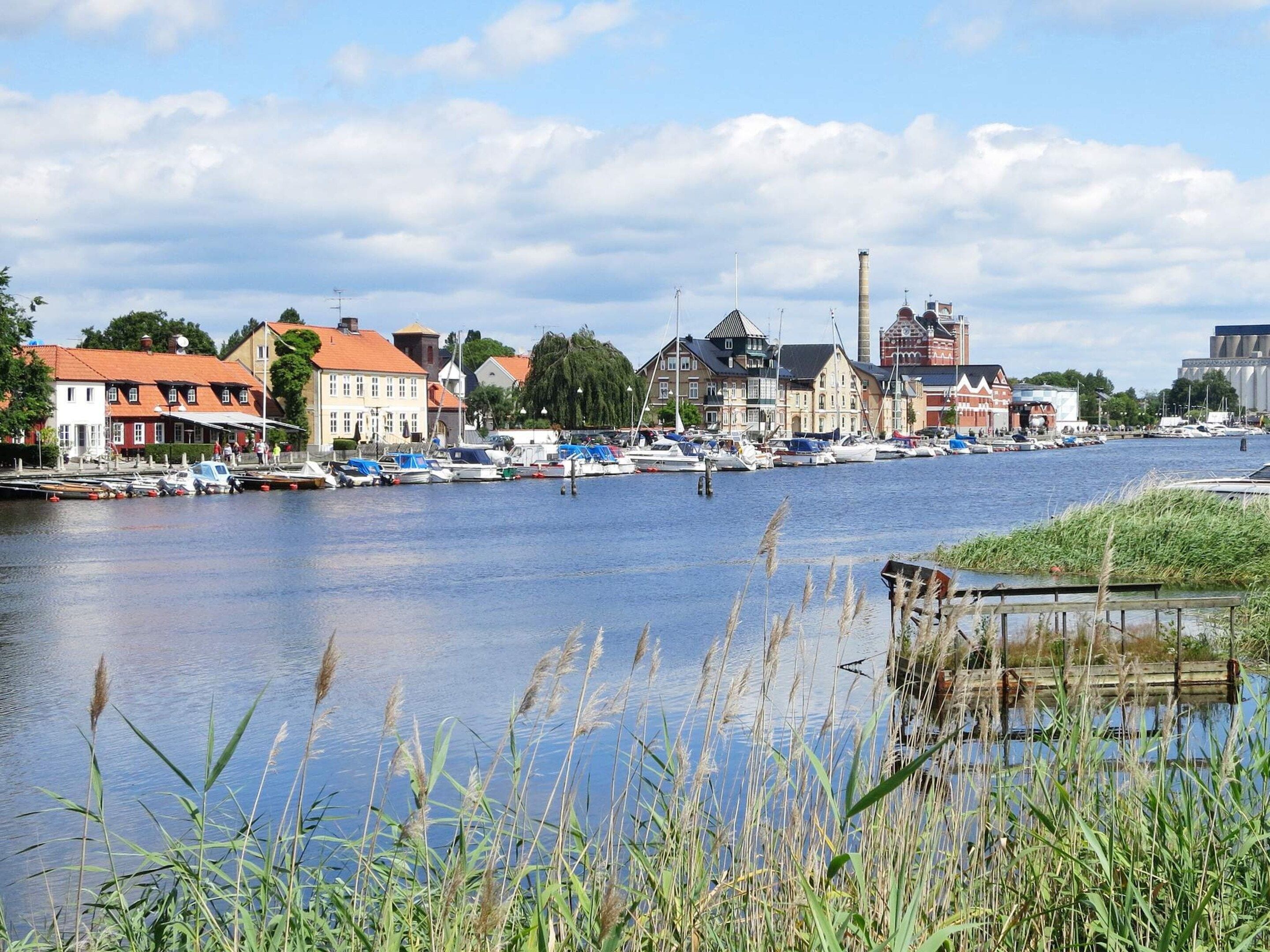 A riverside town with boats docked along the water, historic buildings, and a bridge in the background.