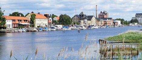 A riverside town with boats docked along the water, historic buildings, and a bridge in the background.