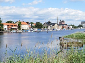 A riverside town with boats docked along the water, historic buildings, and a bridge in the background.