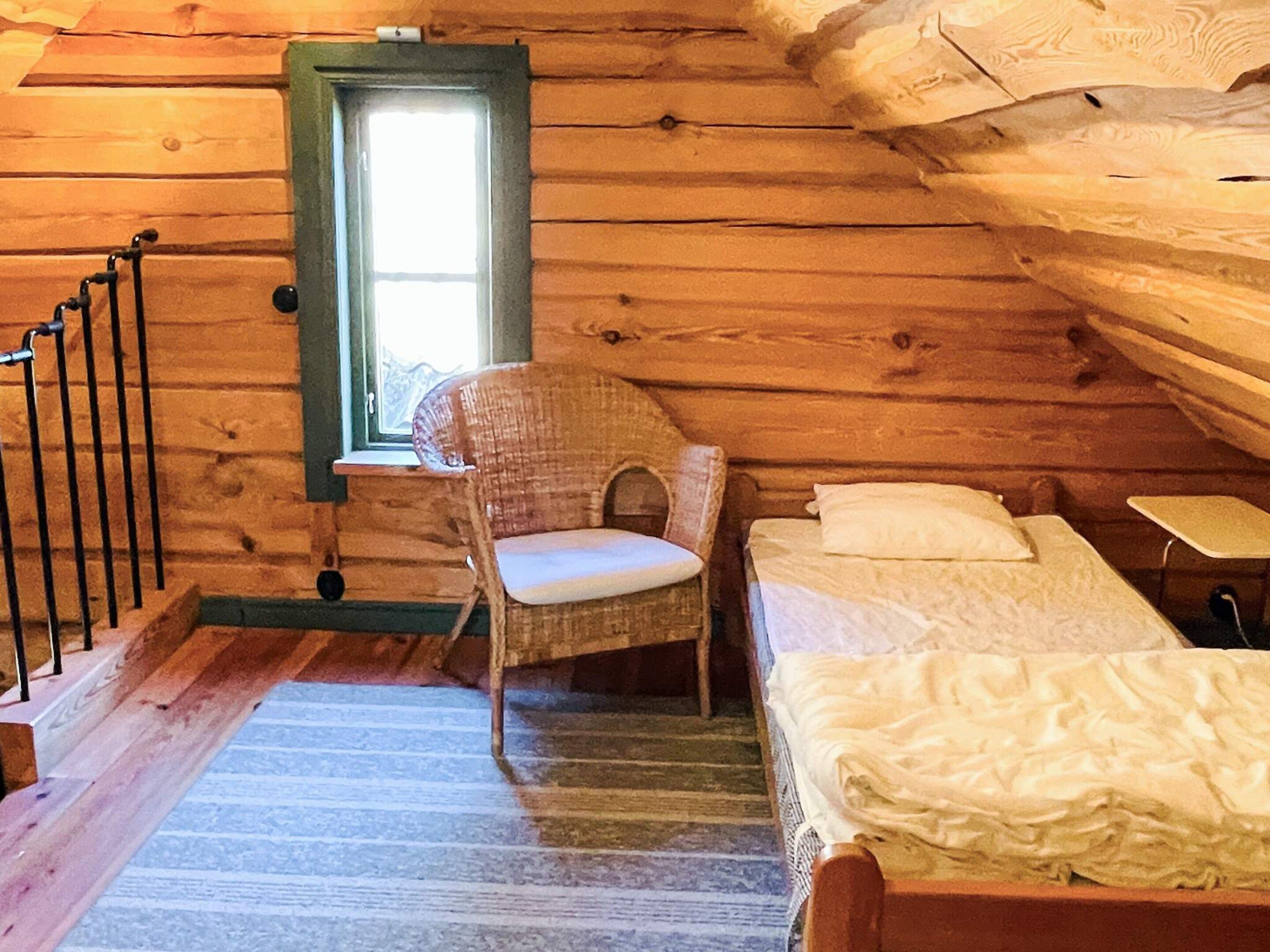 Una habitación tipo cabaña de madera con una cama, una silla, una ventana y una escalera.