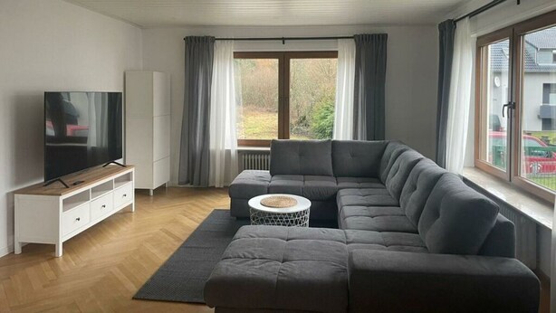 A modern living room with a grey sectional sofa, a TV on a wooden stand, a white cabinet, and a large window with curtains. - Holiday Apartment at Schillerfelsen (Dahn)