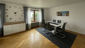 A modern room with a wooden floor, a white table with chairs, a clock on the wall, and a window with curtains. - Holiday Apartment at Schillerfelsen (Dahn)