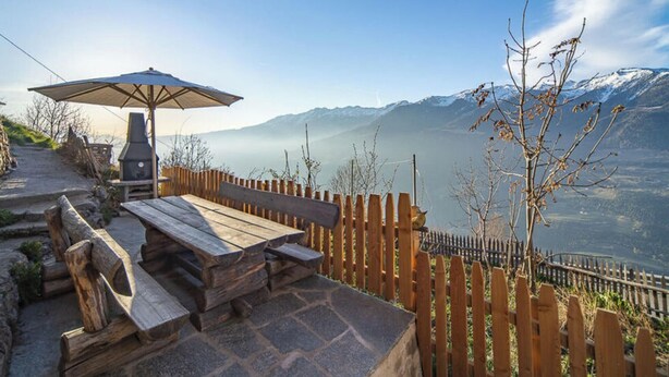 A patio with a wooden table and bench, a barbecue grill, and a wooden fence overlooking a mountainous landscape.