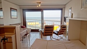 A hotel room with a bed, a desk, a chair, and a view of the beach and pier through the large window. - Family Apartment in Nieuwpoort (Nieuwpoort)