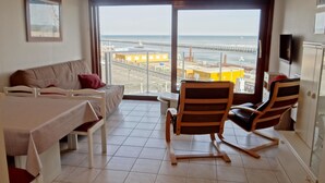A hotel room with a bed, a sofa, a dining table, two chairs, and a large window offering a view of the beach and pier. - Family Apartment in Nieuwpoort (Nieuwpoort)