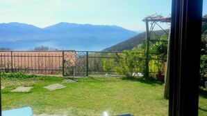 Vista desde una ventana con un jardín césped, una cerca metálica, un muro de ladrillo y montañas al fondo.