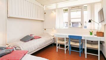 A small, compact room with a single bed, a desk, and a chair. There is a window with curtains, a lamp on the desk, and a small table with a basket and pens.
