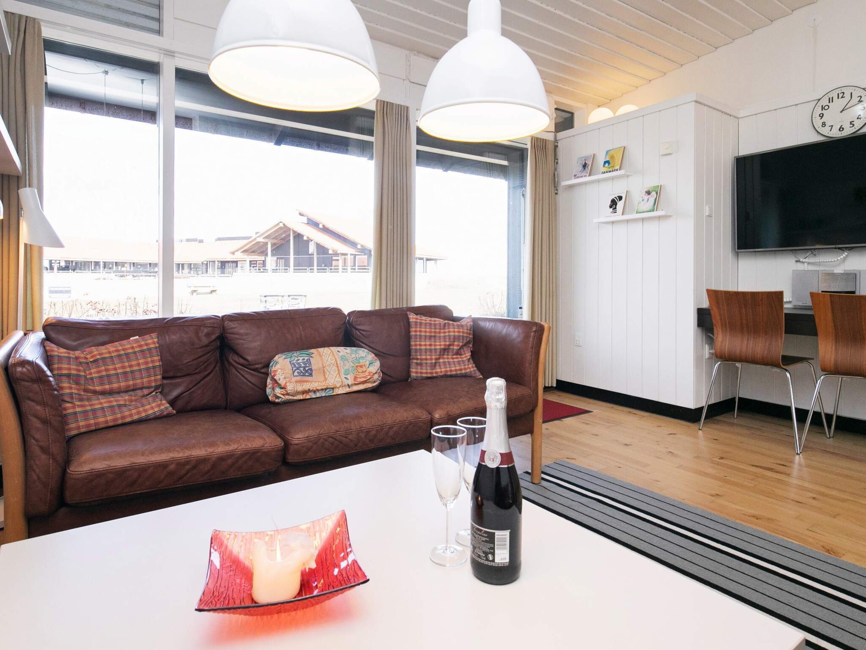 A modern living room with a brown leather sofa, a white table with a bottle and glasses, and a large window with a view of a building outside.
