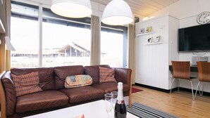 Una sala moderna con un sofá de cuero marrón, una mesa blanca con una botella y copas, y un ventanal con vistas a un edificio.