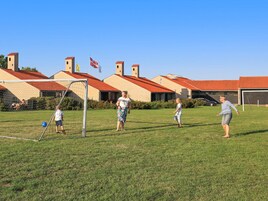 Un campo cubierto de hierba con un arco de fútbol, edificios con tejados rojos y banderas al fondo.