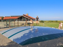 Un patio de juegos con un trampolín y estructuras para escalar, frente a un edificio de techo rojo.