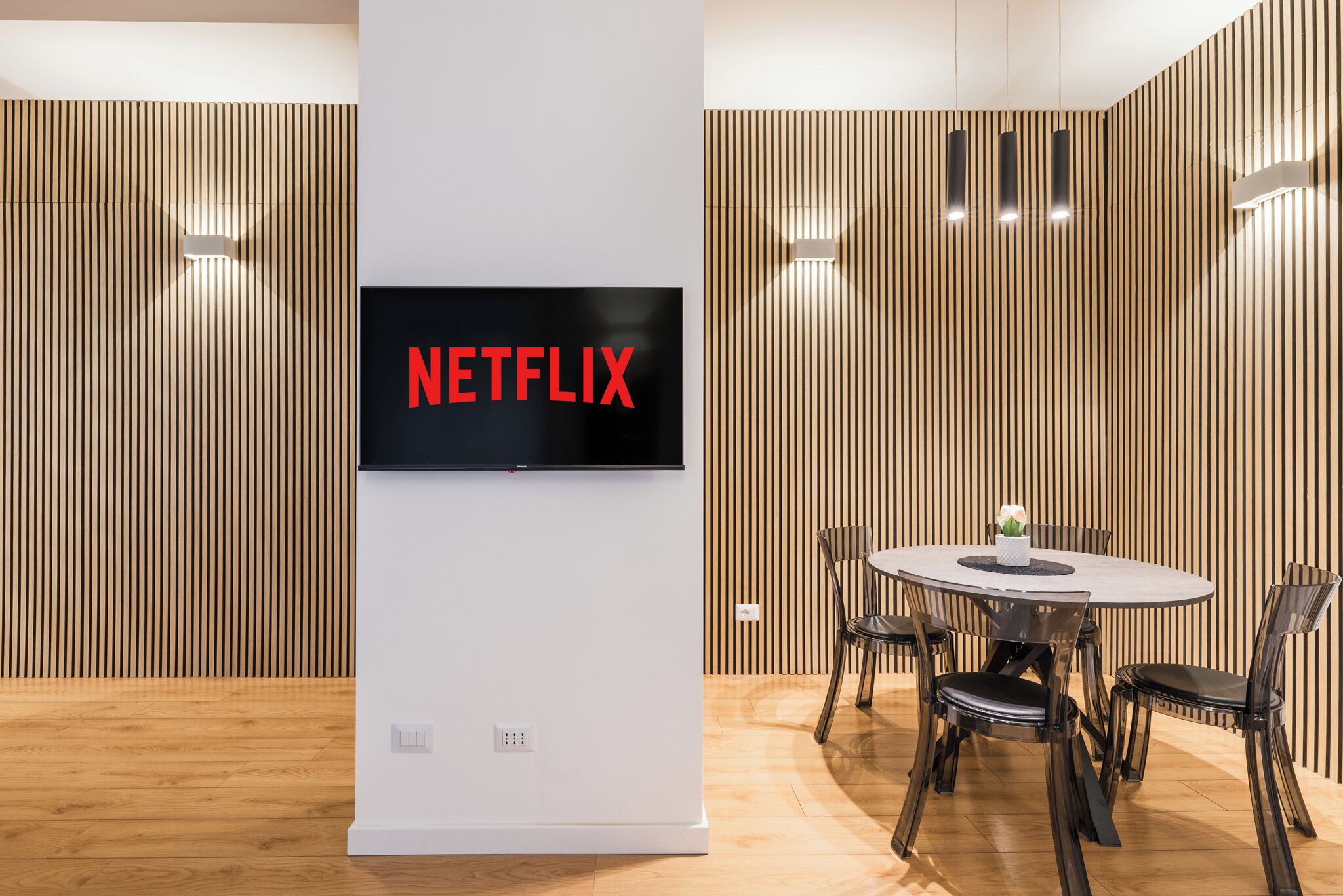 Un intérieur moderne avec un téléviseur fixé au mur affichant le logo « NETFLIX » sur un mur blanc, une table ronde avec des chaises et un lambris en bois sur les murs.