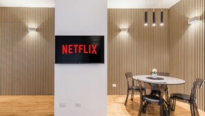 A modern interior with a mounted TV displaying "NETFLIX" on a white wall, a round table with chairs, and wooden paneling on the walls. - THE FIFTH PROPERTIES ROME (Roma)